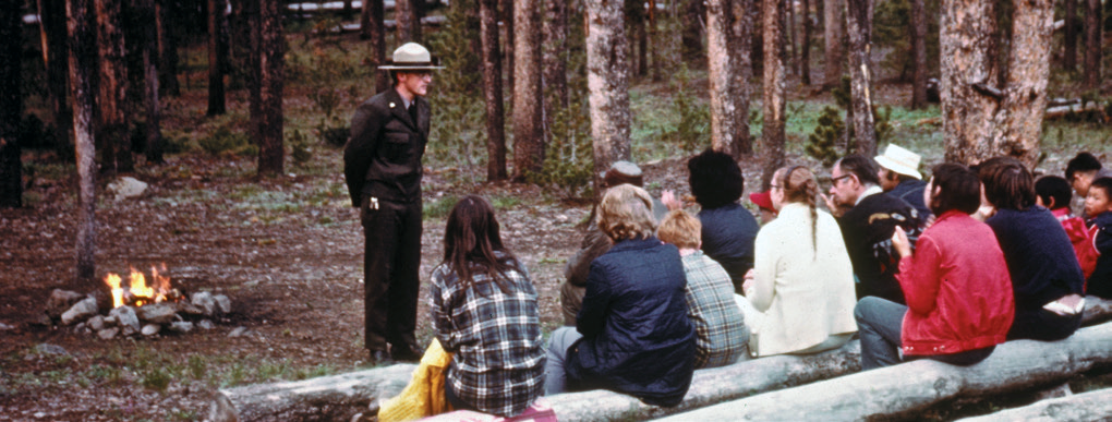 A young man standing in front of an audience with a campfire in the background of a campground.