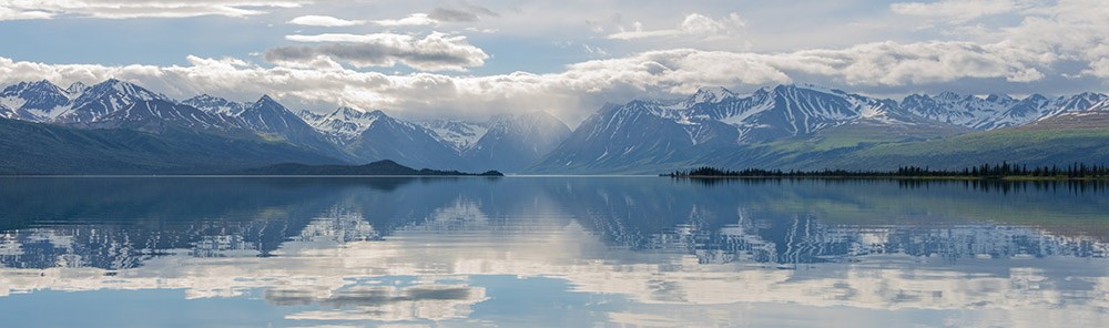 A panorama view of Telaquana Lake with sky and clouds reflecting on the calm lake surface.