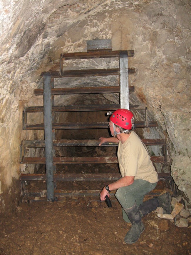 A man kneeling before a boarded up cave entrance with a red hard hat