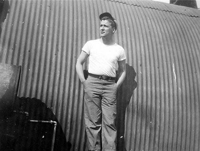 Black and white photo of young man standing in front of rounded, corrugated metal building.