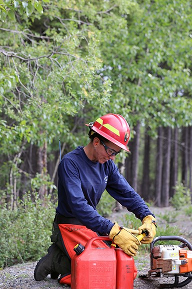 A female firefighter leans over her chainsaw to do a maintenance check and to fill it with gas and bar oil.