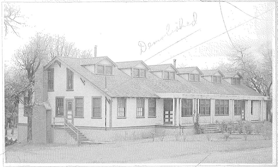 Black and white photo of a rectangular building with chimney, windows, and a porch.