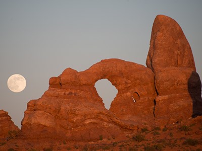 the full moon rises above a reddish stone arch