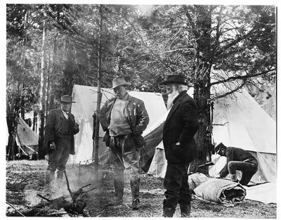 Black and white photo of Teddy Roosevelt standing with other men around a smoldering pile of branches
