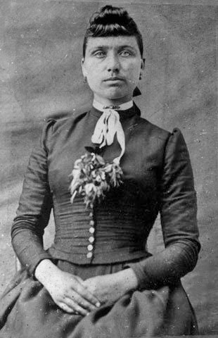 A black and white, formal portrait from the 1880s of a young woman is a high collared, dark dress with a small bouquet of flowers tucked into the bodice.