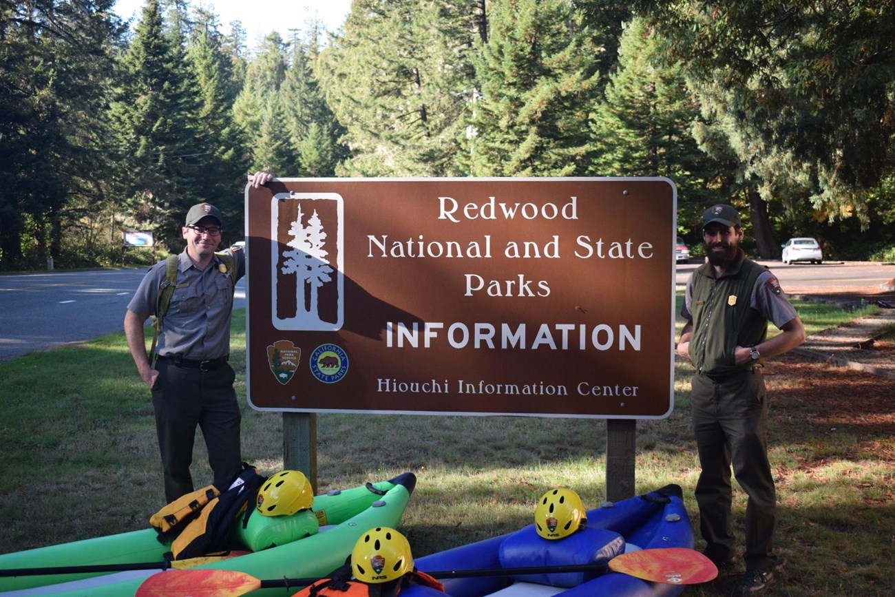 two park rangers standing by the Redwoods National and State Parks sign with two kayaks in front.