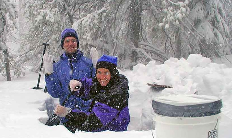 two technicians in colorful winter jackets and hats standing in a snow pit up to their waists, smiling and shoveling snow while snow is falling