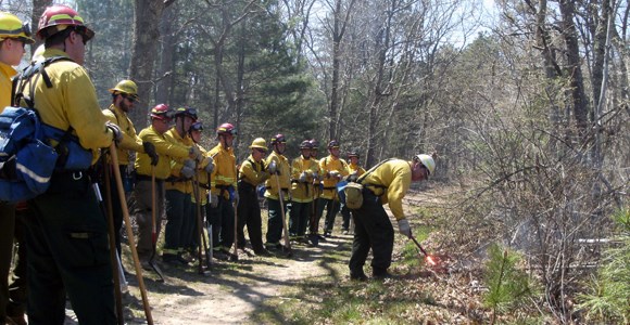 A group of firefighters is gathered while the lead firefighter ignites grass with an ignition device.