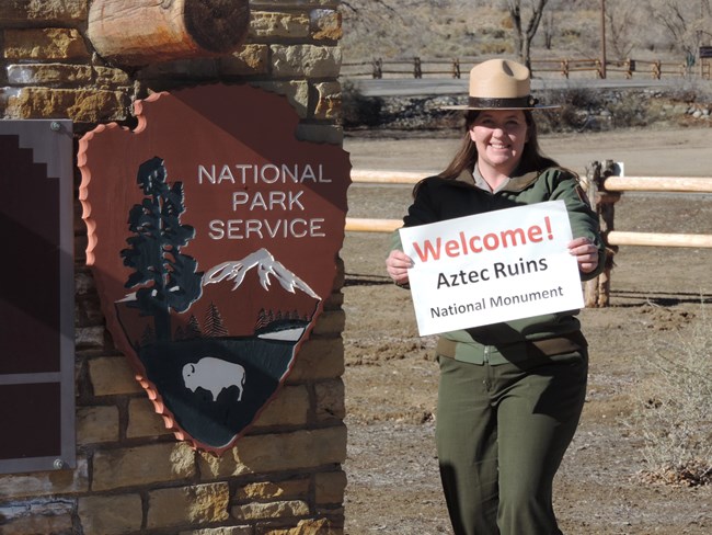 A ranger posing next to a large National Park Service arrowhead holding a sign that says "Welcome! Aztec Ruins National Monument."