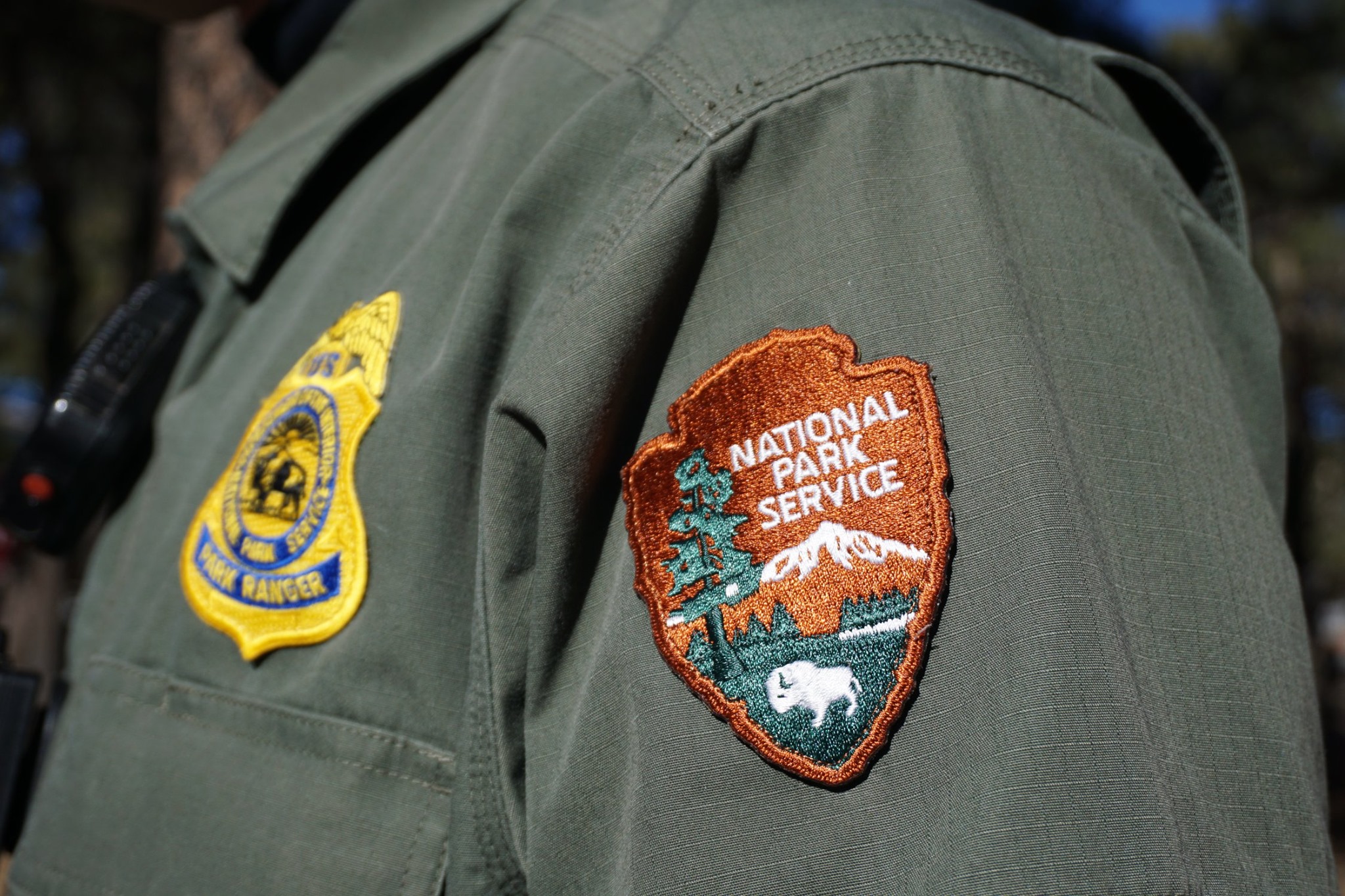 An embroidered orange and green arrowhead patch with a white bison and the words National Park Service at the top. It is on a green uniform shirt with a yellow and blue embroided Park Ranger badge.