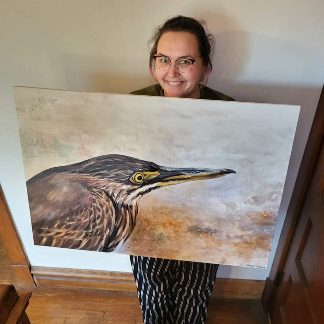 Lindsey Winslow holds one of her finished paintings in her studio. The canvas painting depicts a bird on a foggy background with brown and white feathers, gold under beak and black top beak and golden eyes.