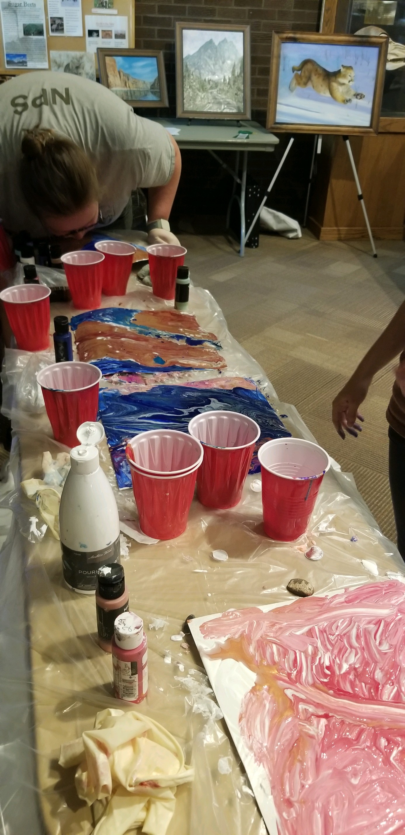 A table covered in plastic and red cups is used to pour paint on to paper to make art work using layers of paint.