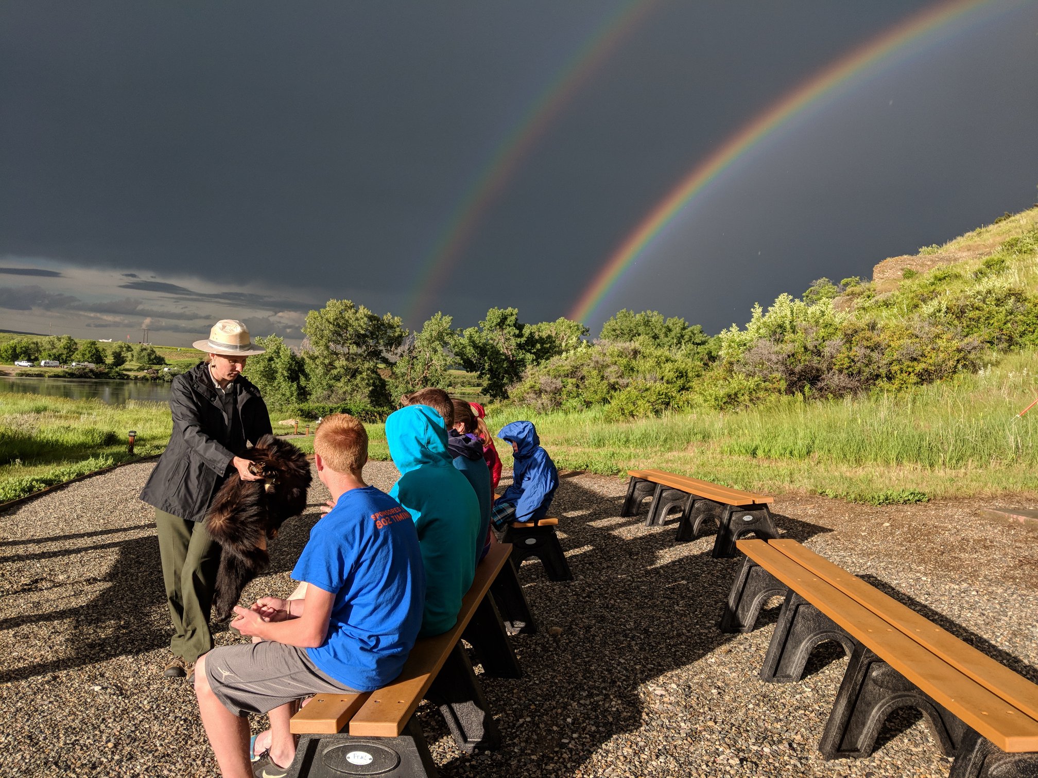 A ranger holds out an animal pelt for children to touch. A passed storm and rainbows are in the background.