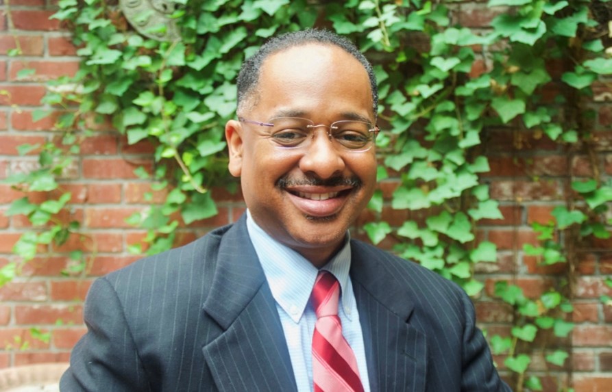 A well dressed man in a suit and tie smiles at the camera in front of a brick wall covered in green ivy.