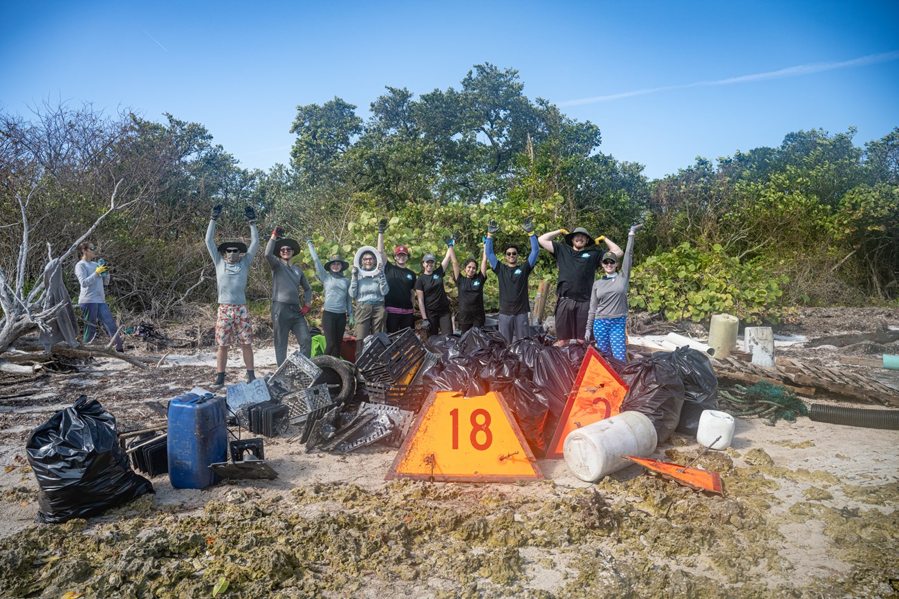 A group of park staff and volunteers stand on a beach surrounded by several trash bags, crates, and various other types of marine debris including a large triangular channel marker with the number 18 printed on it.