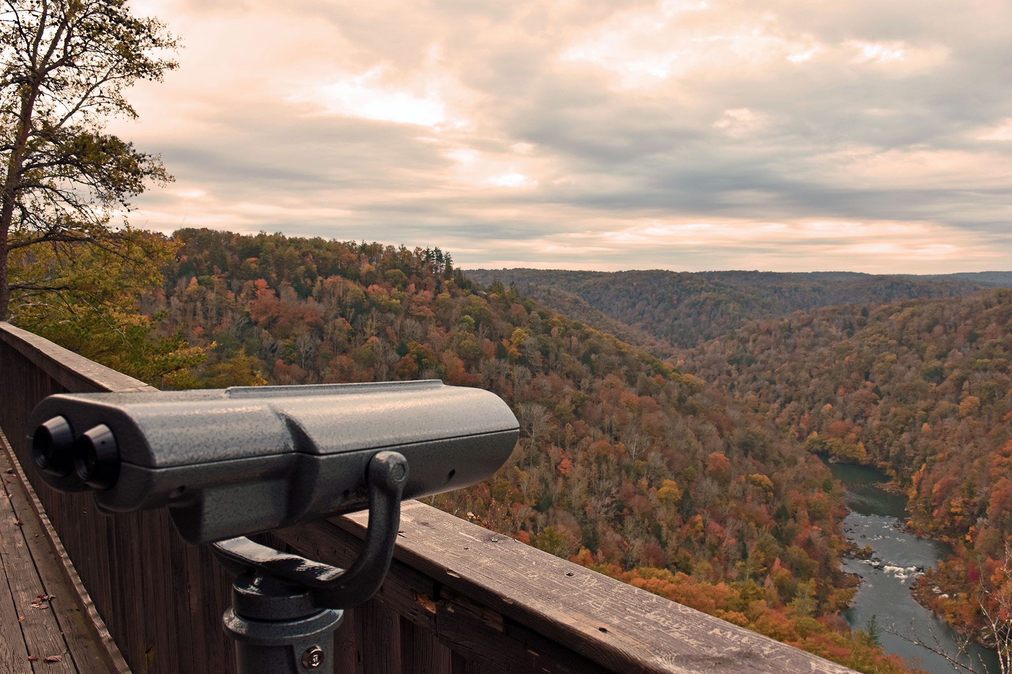 A scenic view from the East Rim Overlook showing a valley of autumn-colored trees and a river winding below, with a public telescope in the foreground on a wooden observation deck.