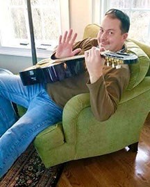 A musician playing the guitar on a green chair waving.