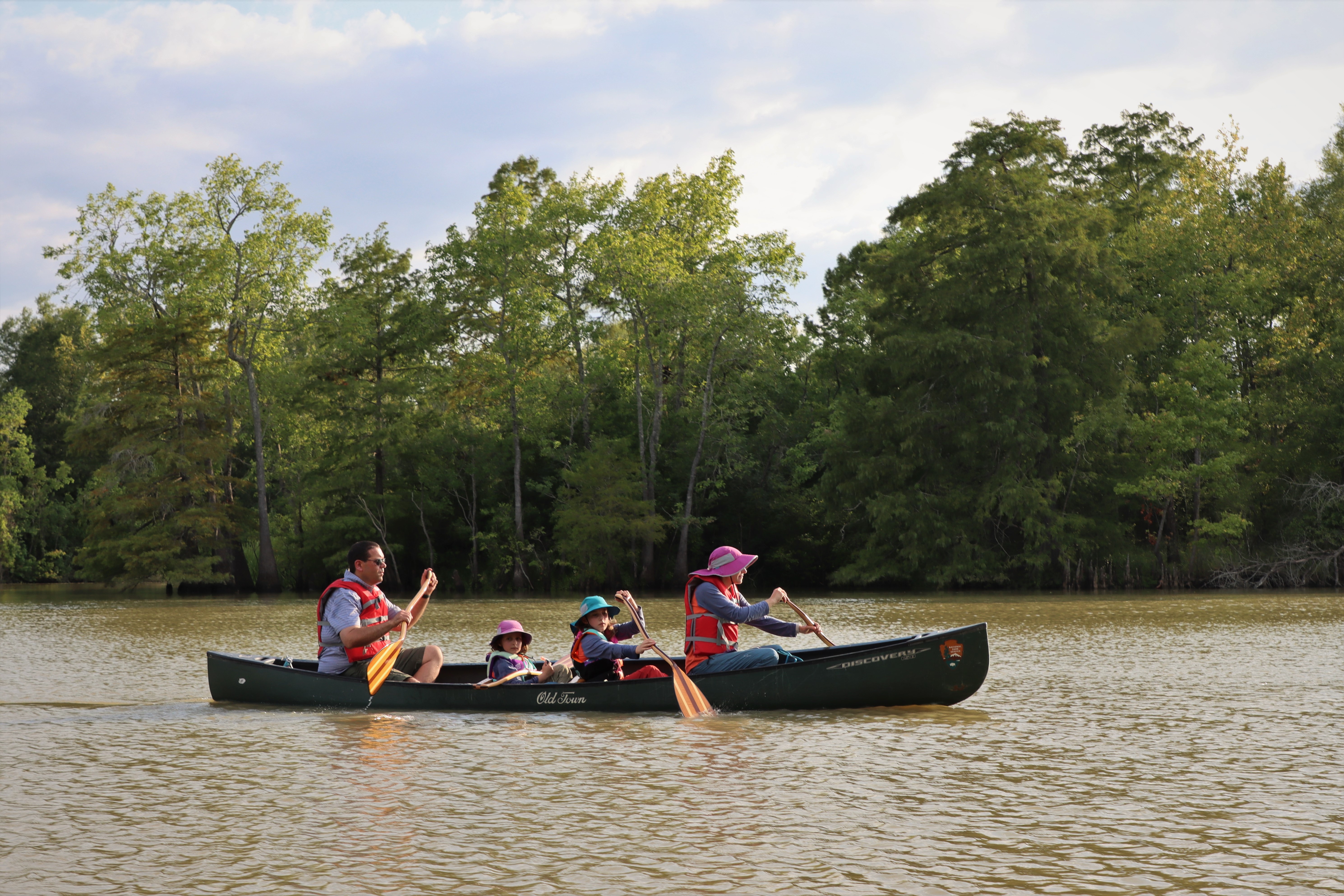 a father, mother, and 2 kids paddling a large green canoe on a wide, forested river.