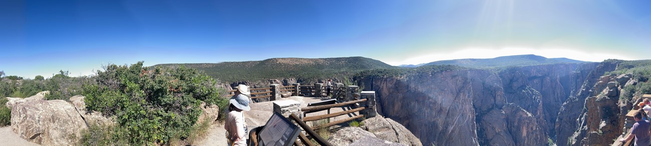 Panoramic image of people standing at a canyon overlook. There are waysides and wooden fencing along the edge of the overlook.