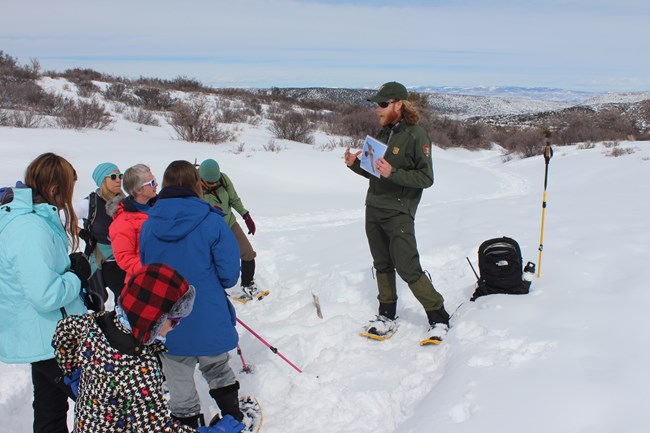 A park ranger in a green uniform holds up a photo to show a group of visitors standing in the snow