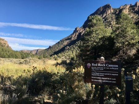 Red Rock Canyon trailhead sign with green shrubs and some rocky cliffs