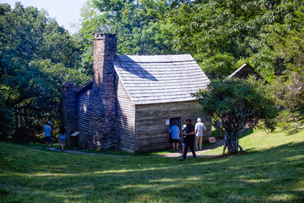 Visitors gather around an older brick and lumber building in a shady grove