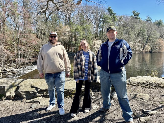 three people standing in front of a forest and a pond