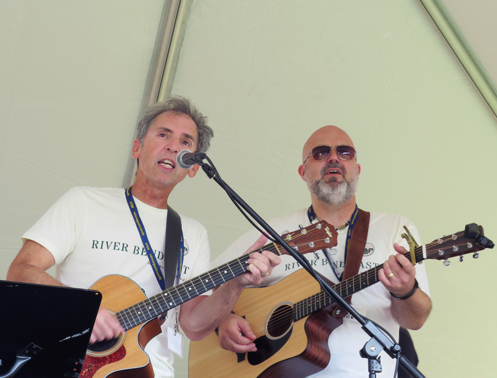 Two men singing while holding guitars under a tent