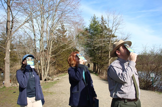 three people holding eclipse glasses look up at the sky.