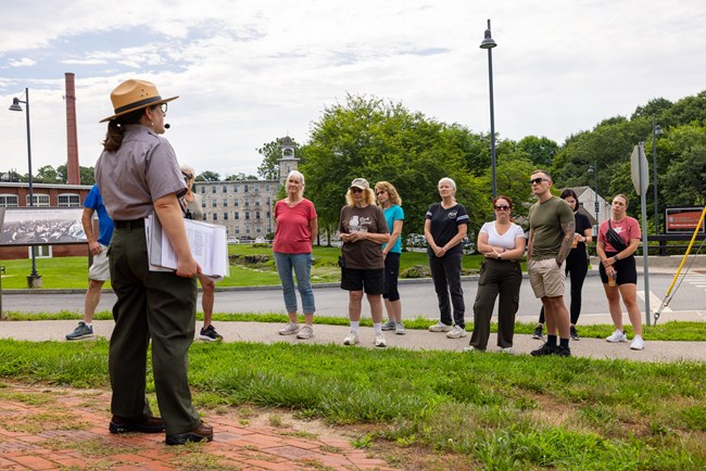 a ranger gives a tour on green grass to a group of people in front of a mill