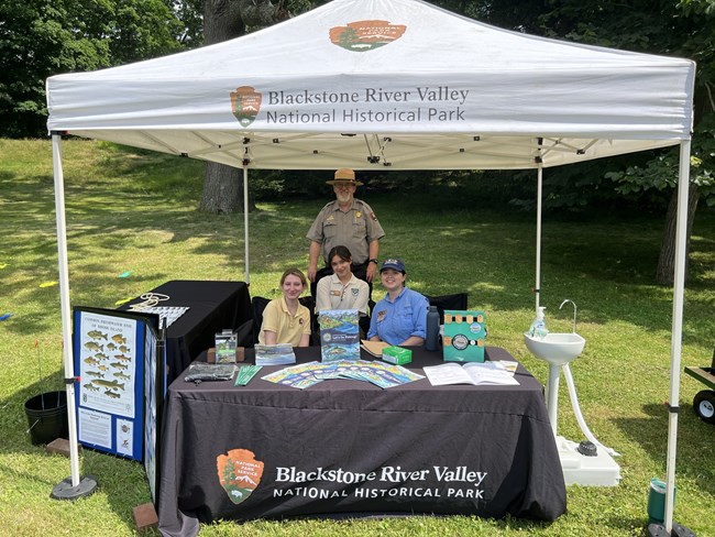 A uniformed park ranger stands behind three interns in polos, surrounded by fishing activities