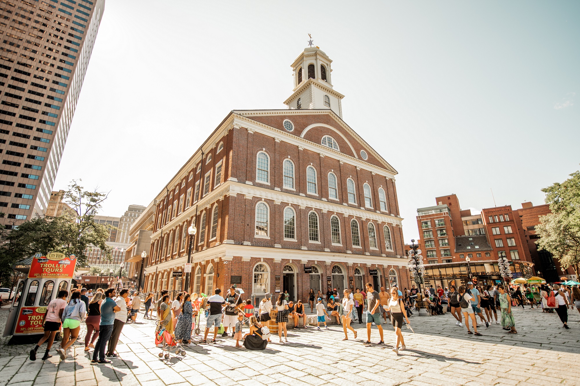 Faneuil Hall, a red brick 3 story building with a cupola. Groups of people gather and walk around the square in front of the building.