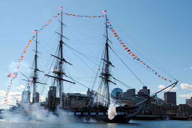 USS CONSTITUTION in Boston Harbor displaying full-dress signal flags draped from bow to stern over her three masts. Signal guns are firing in salute.