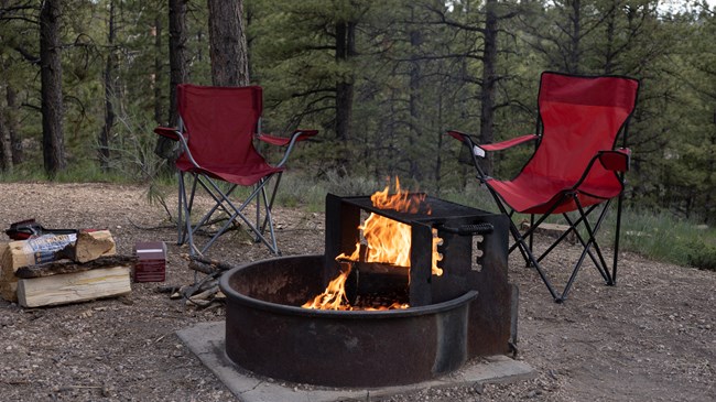 Two empty red chairs sit next to an unattended fire in a fire pit.