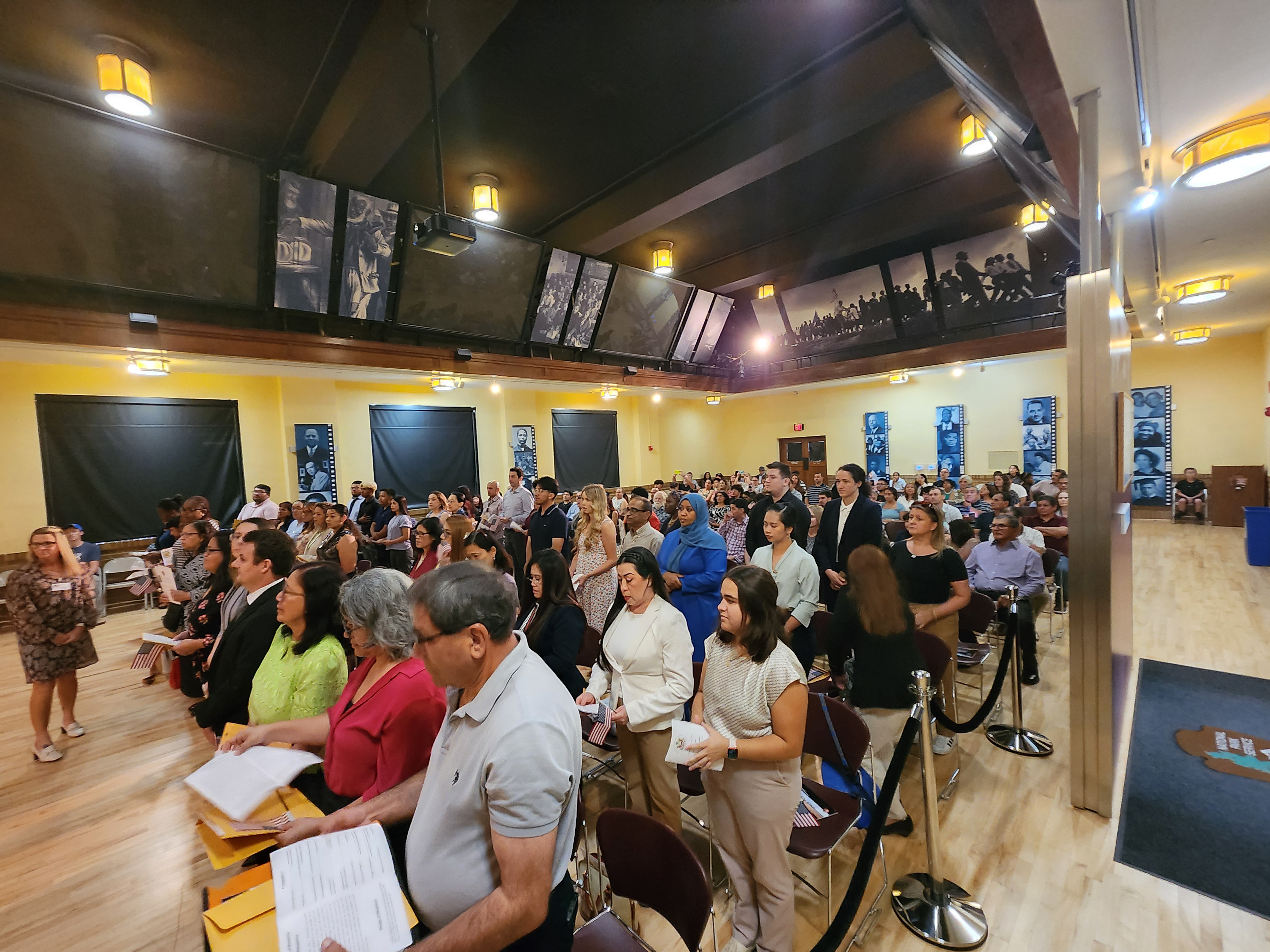 People stand in an auditorium surrounded by friends and family while holding folders of documents.