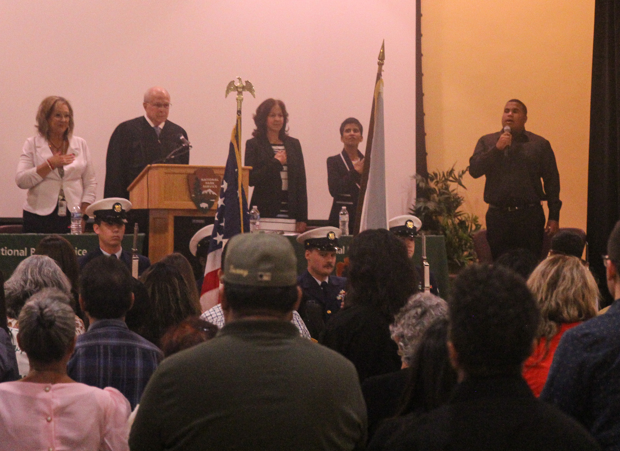 Five people can be seen standing on the top of a stage with their hands over their heart pledging allegiance to the United States flag. A large crowd can be seen looking towards the stage and pledging allegiance to the United States flag.