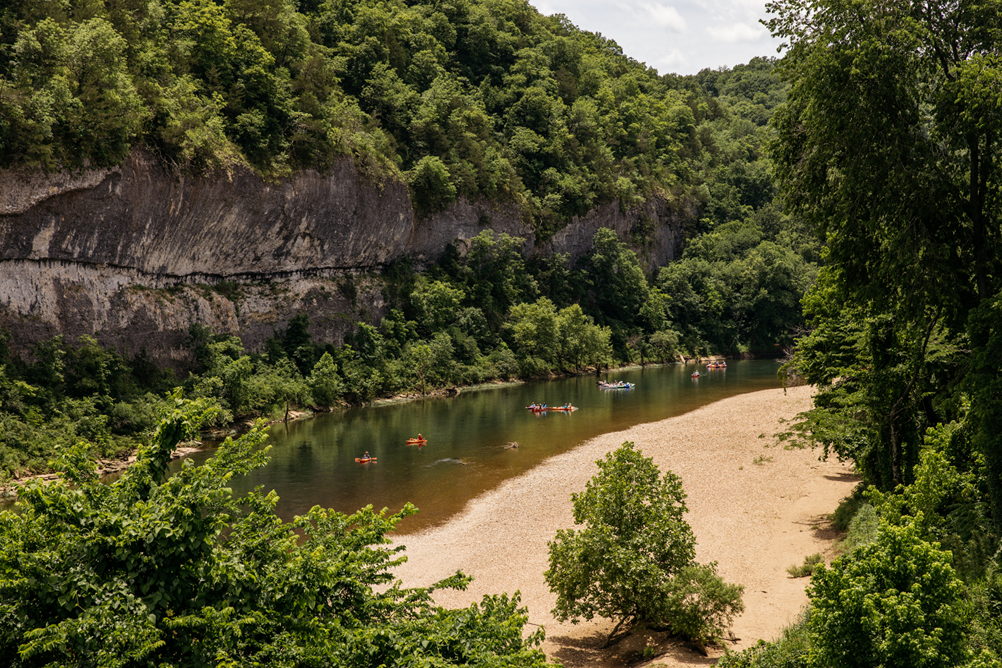 A stretch of river, with several colorful kayaks on it, can be seen flowing at the base of a tan and gray streaked bluff. Tree covered hillsides can be seen flanking the far side of the river. A sandy gravel bar stretches the entire length of the river on