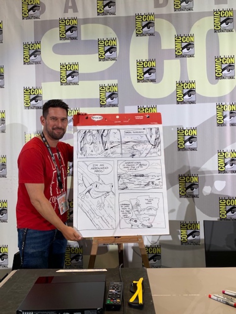 Alonso Nunez from Little Fish Comic Book studio poses next to an easel and large pad of paper with his live-draw, one-page comic on it. The sharpie-drawn comic images depict the Amazon Rainforest and an Arrow Frog.