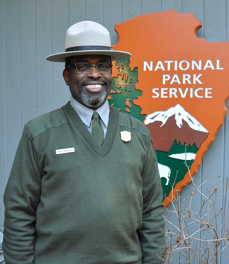 Uniformed ranger stands infront of the National Park Service Arrowhead