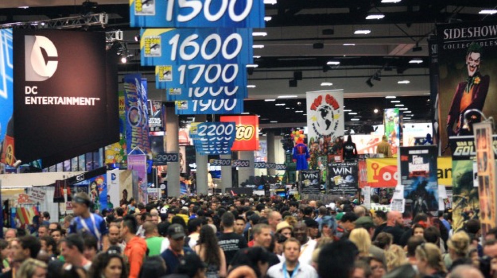 A shot inside the exhibit hall of San Diego Comic-Con 2019. A massive crowd moves through exhibit booths from DC Entertainment, Lego, Top Cow, Sideshow Collectibles, and others.