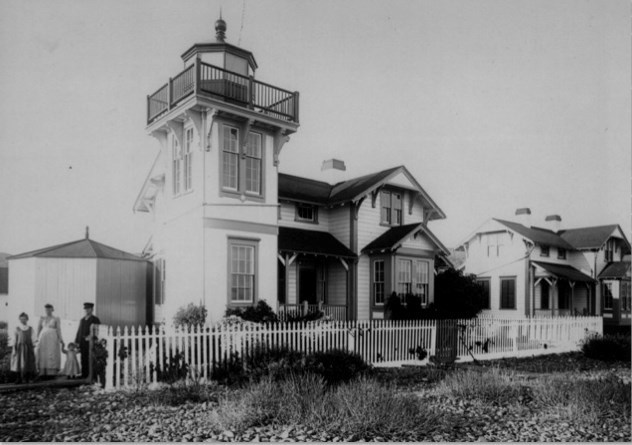 Black and white photograph of Ballast Point Lighthouse, featuring a square tower attached to a clapboard Mission Revival-style house. Several people, including a lighthouse keeper and his family, stand near a white picket fence surrounding the lighthouse.