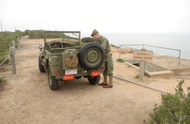 A reenactor in WWII military attire stands next to a green military jeep. The jeep is parked near a sign reading "Battery H Control Station," with the Pacific Ocean in the background and coastal shrubbery on the left.