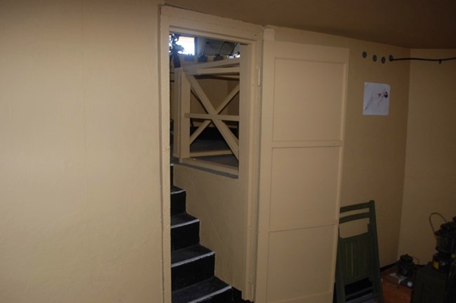 A view from the lower bunker looking up at the restored stairwell and gate. The original staircase leads to the upper level, with the wooden gate visible at the top. The space has been restored to maintain its WWII-era appearance.