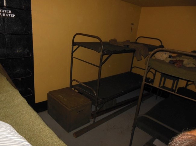 A WWII-era bunkroom with metal-framed bunk beds stacked two high and a storage chest on the floor. The room is sparse and utilitarian, reflecting the living conditions of soldiers stationed in the bunker during wartime.