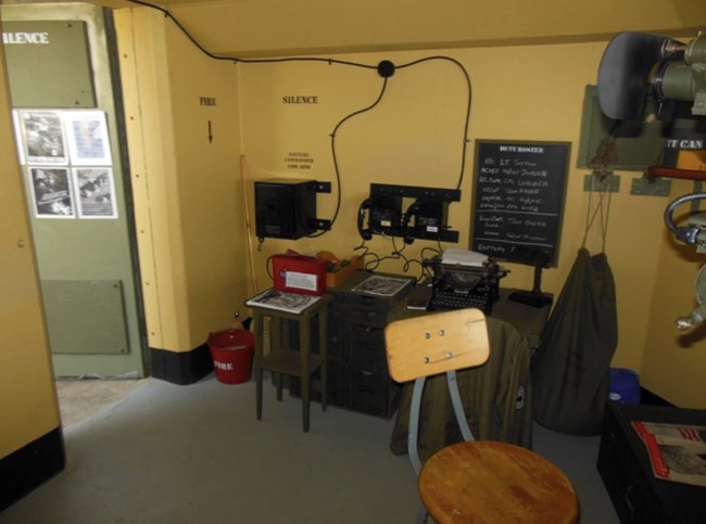 A WWII-era communication room inside the Battery Commander's station, featuring a desk with a typewriter, radio equipment, and a chalkboard with messages. "Fire" and "Silence" commands are painted on the walls, and a stool sits in front of the desk.