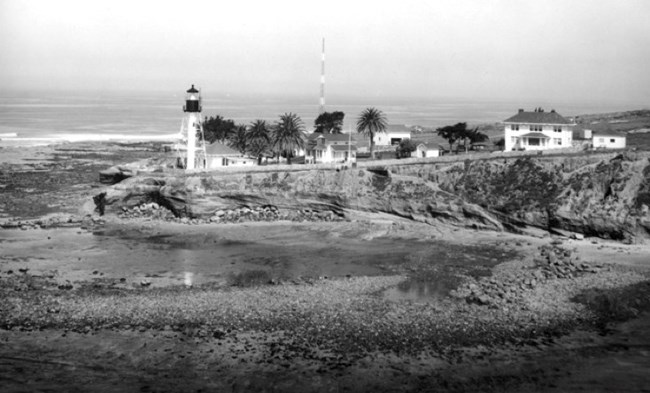 Black and white photograph of the Point Loma Light Station, featuring the lighthouse, several buildings, and palm trees on a rocky coastline. The ocean and a tall antenna are visible in the background.