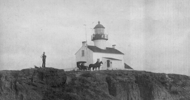 Black and white photograph of Point Loma Lighthouse in 1890, situated on a cliff. A person stands beside a horse-drawn carriage in front of the lighthouse, which features a central tower and attached dwelling.