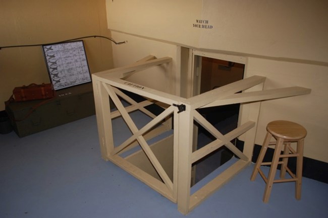 A stairwell inside the lower bunker with a "Watch Your Head" sign above. A ship silhouette chart is positioned nearby, used for target identification. A wooden stool sits next to the stairwell, emphasizing the compact nature of the space.