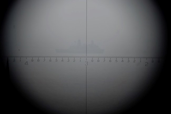 The view through an M1910A1 azimuth scope, showing crosshairs and distance markings. A ship is faintly visible in the foggy distance, illustrating the scope's magnification and use in tracking targets.