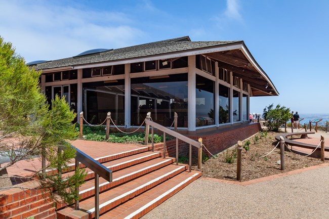 Exterior view of the Cabrillo National Monument Visitor Center, featuring large glass windows and a slanted roof, with steps leading up to the entrance surrounded by greenery and a brick walkway.